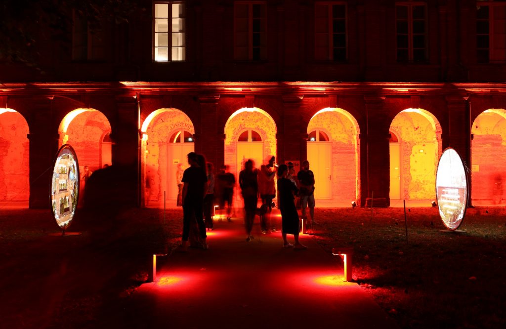 personnes sur un cheminement regardant des vidéoprojection en extérieur sur des toiles tendus avec éclairage d'ambiance en rouge