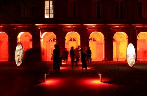 personnes sur un cheminement regardant des vidéoprojection en extérieur sur des toiles tendus avec éclairage d'ambiance en rouge
